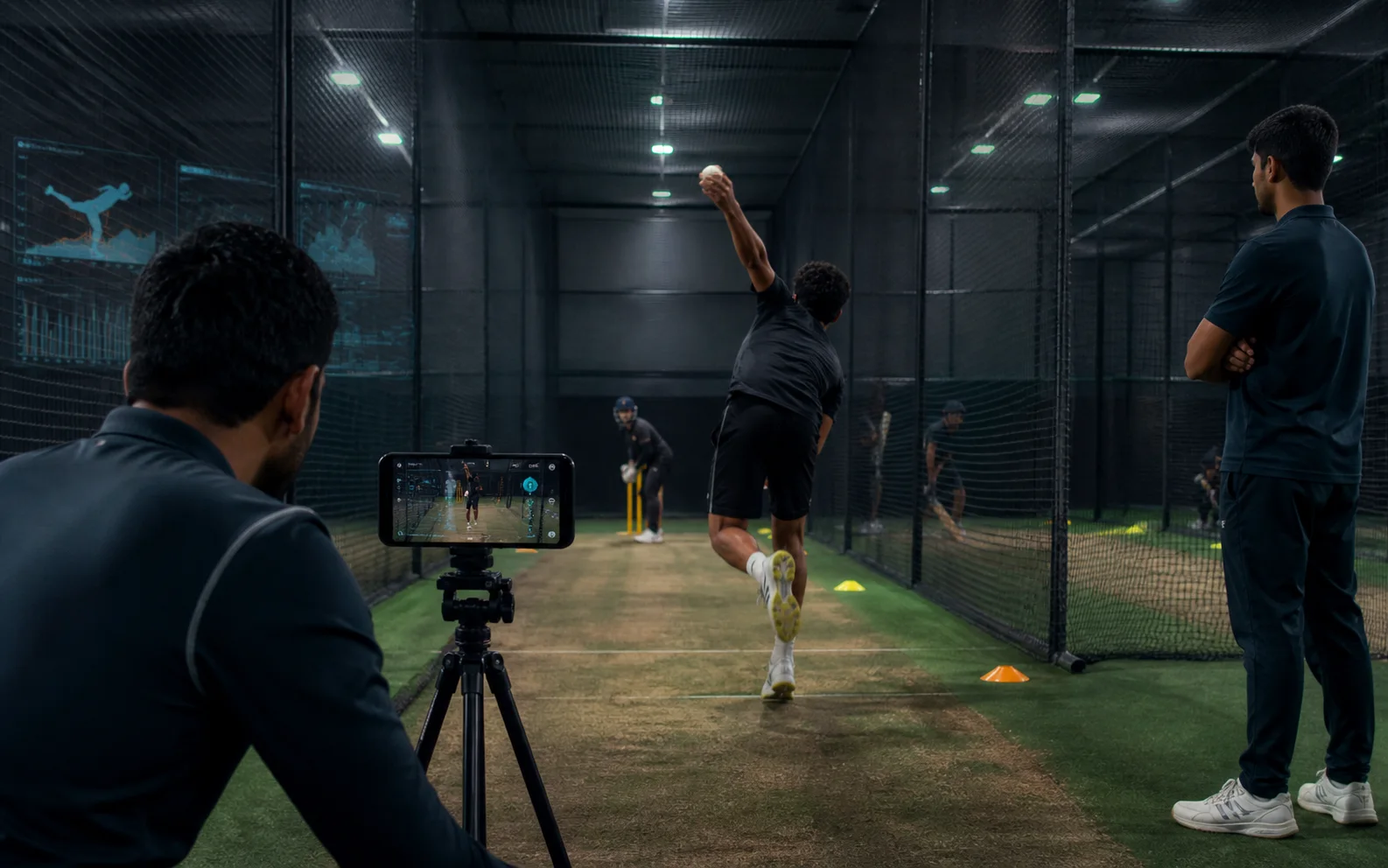 Coach records an indoor cricket net session on a smartphone tripod while academy staff watch the delivery